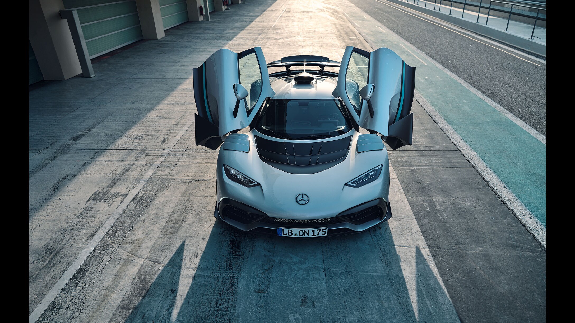 Obrázok zachytáva čelný pohľad na strieborný hyperautomobil Mercedes-AMG ONE s otvorenými krídlovými dverami. Vozidlo stojí v boxovej uličke pretekárskeho okruhu.