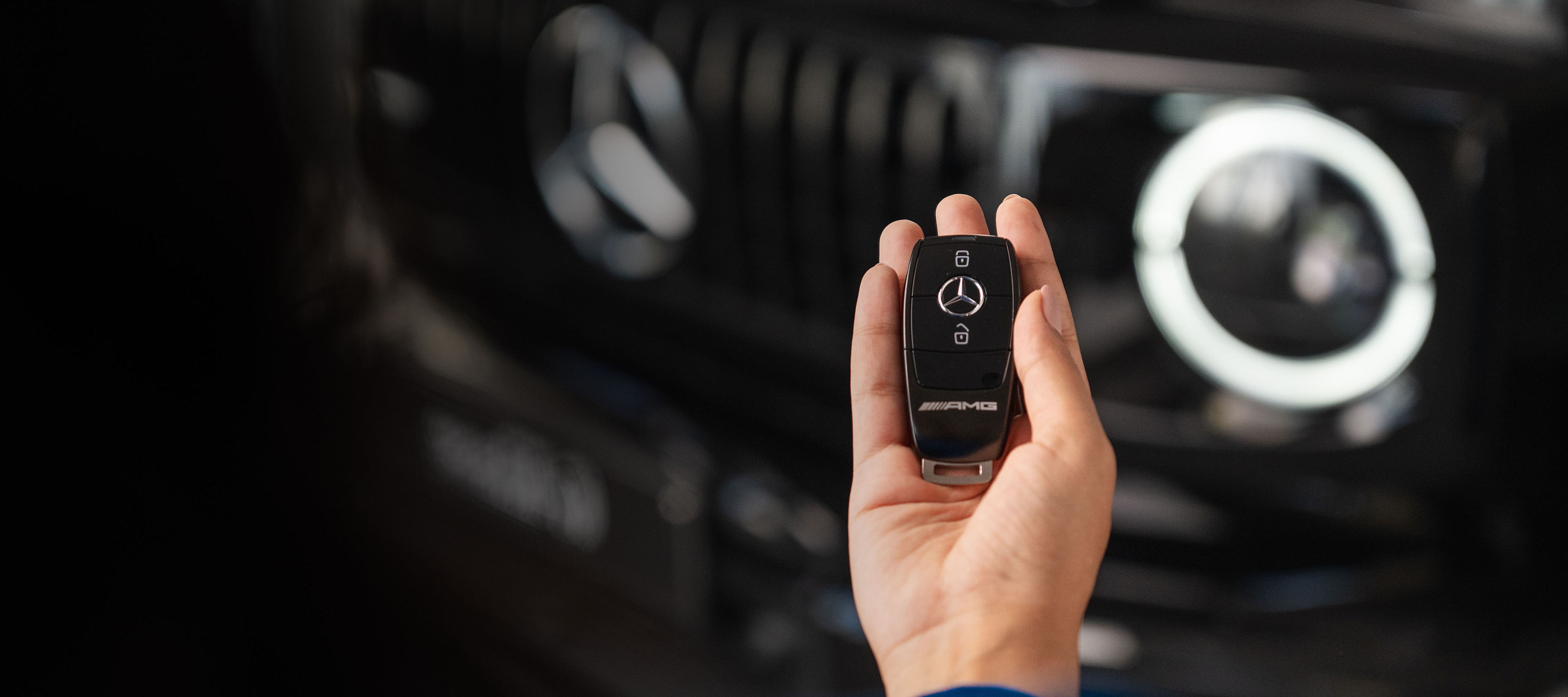 O společnosti Mercedes-Benz Financial Services | Finanční služby | Ruka osoby drží kľúčenku k vozidlu Mercedes AMG, s prednou maskou vozidla v pozadí.