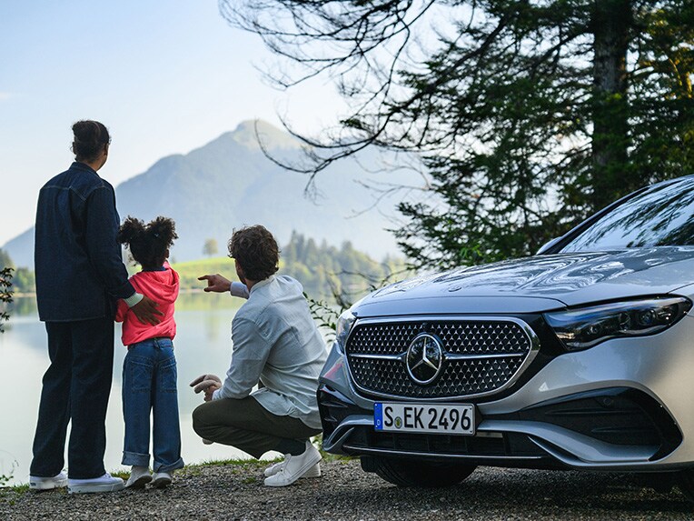 Dvaja ľudia a dieťa sa pozerajú na a hora v diaľke vedľa vozidla Mercedes-Benz.