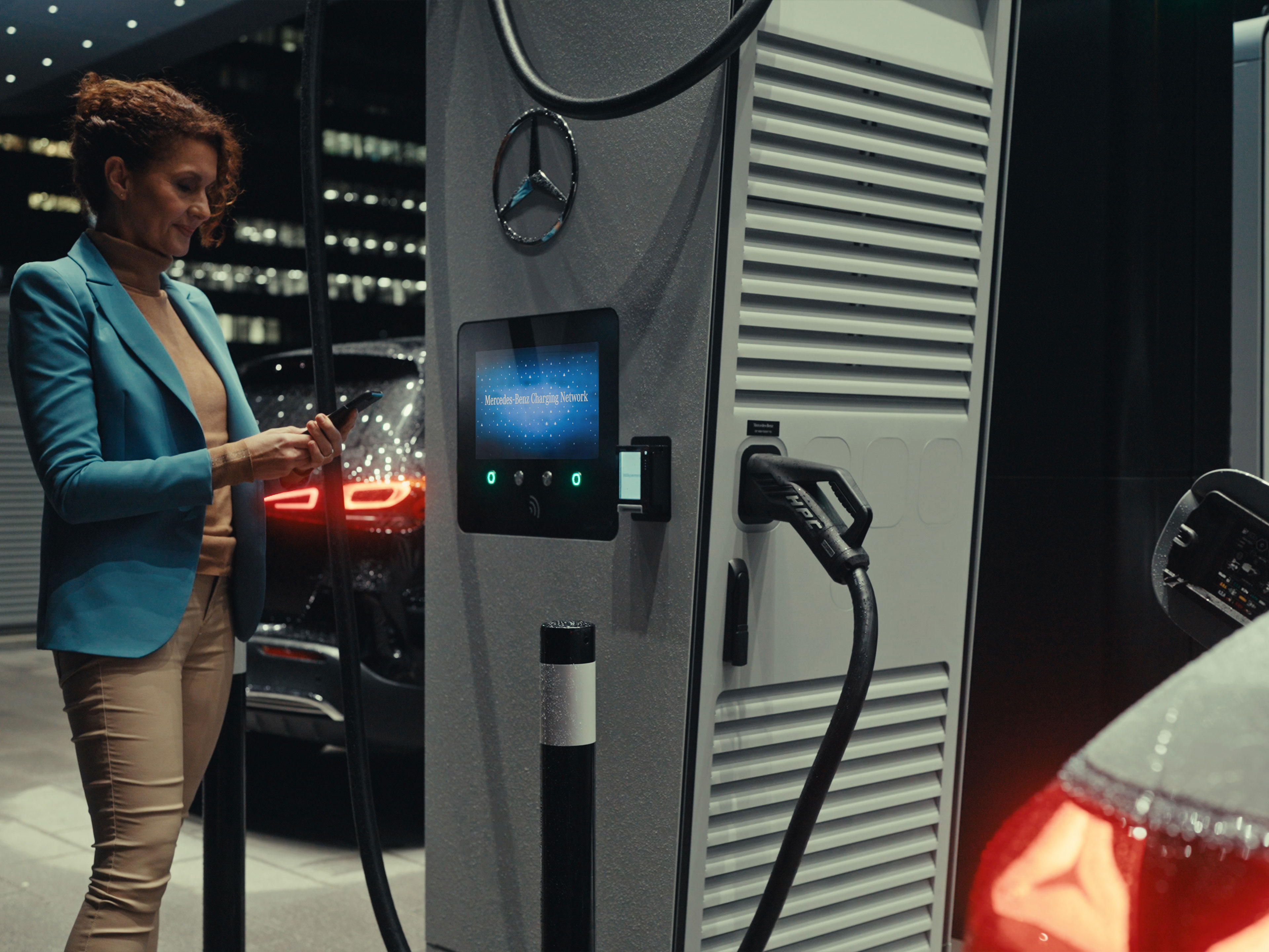 Aplikácia Mercedes-Benz | Nabíjanie na cestách | Mercedes-Benz Žena stojí vedľa nabíjacej stanice s chytrým telefónom v ruke.