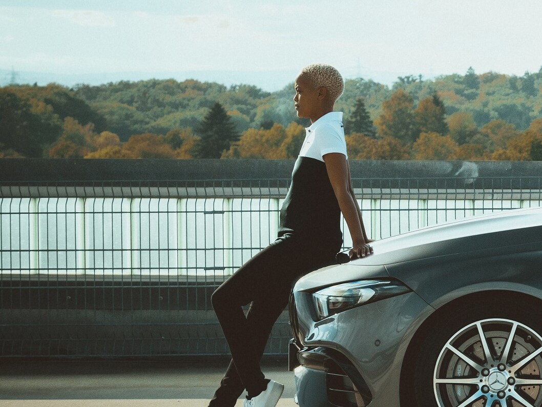 Mladá žena v&nbsp;outfite z&nbsp;kolekcie AMG Racing sa nedbalo opiera o&nbsp;kapotu motora vozidla Mercedes.