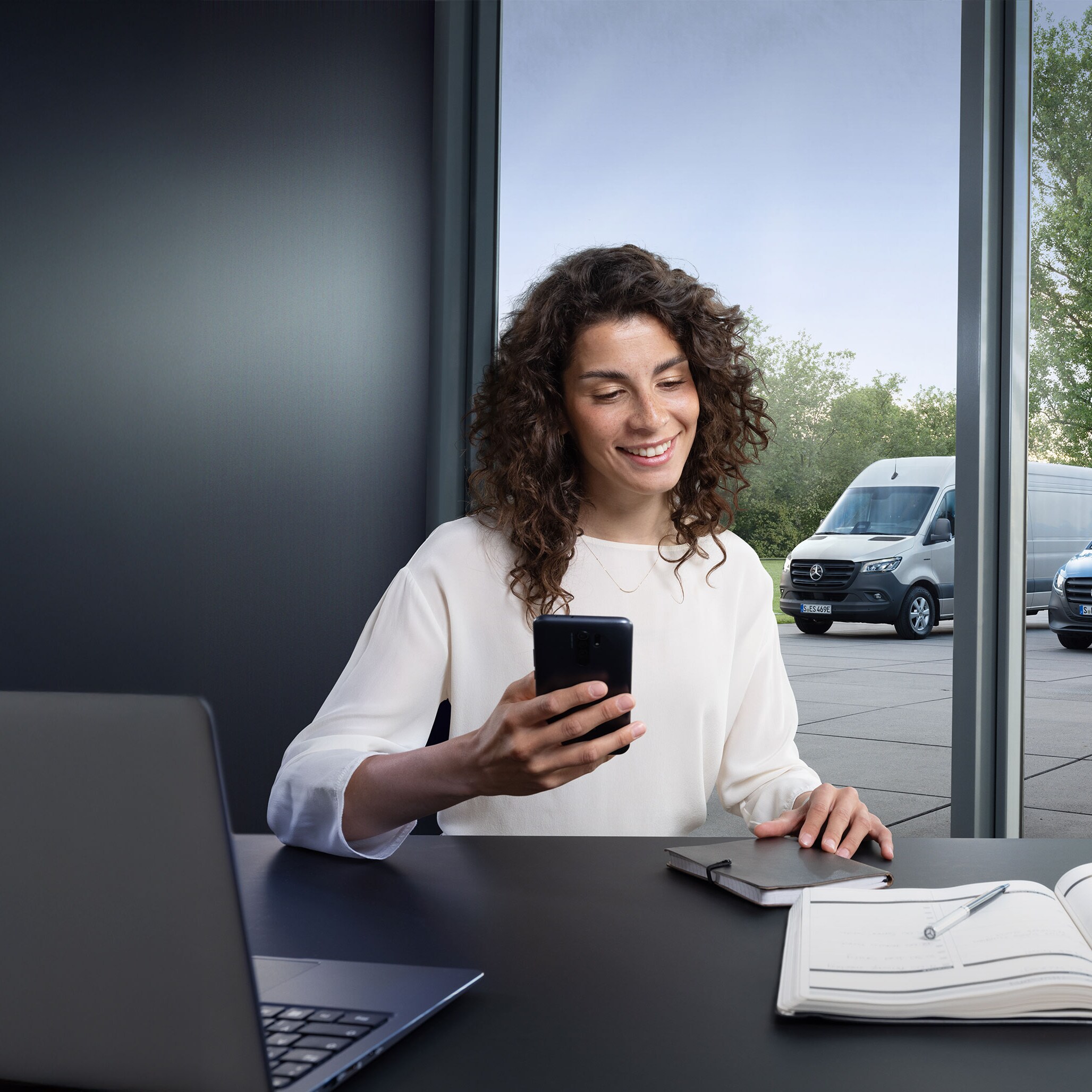 Žena hľadá servisného partnera Mercedes-Benz vo svojom smartfóne, v pozadí je niekoľko úžitkových vozidiel Mercedes-Benz.