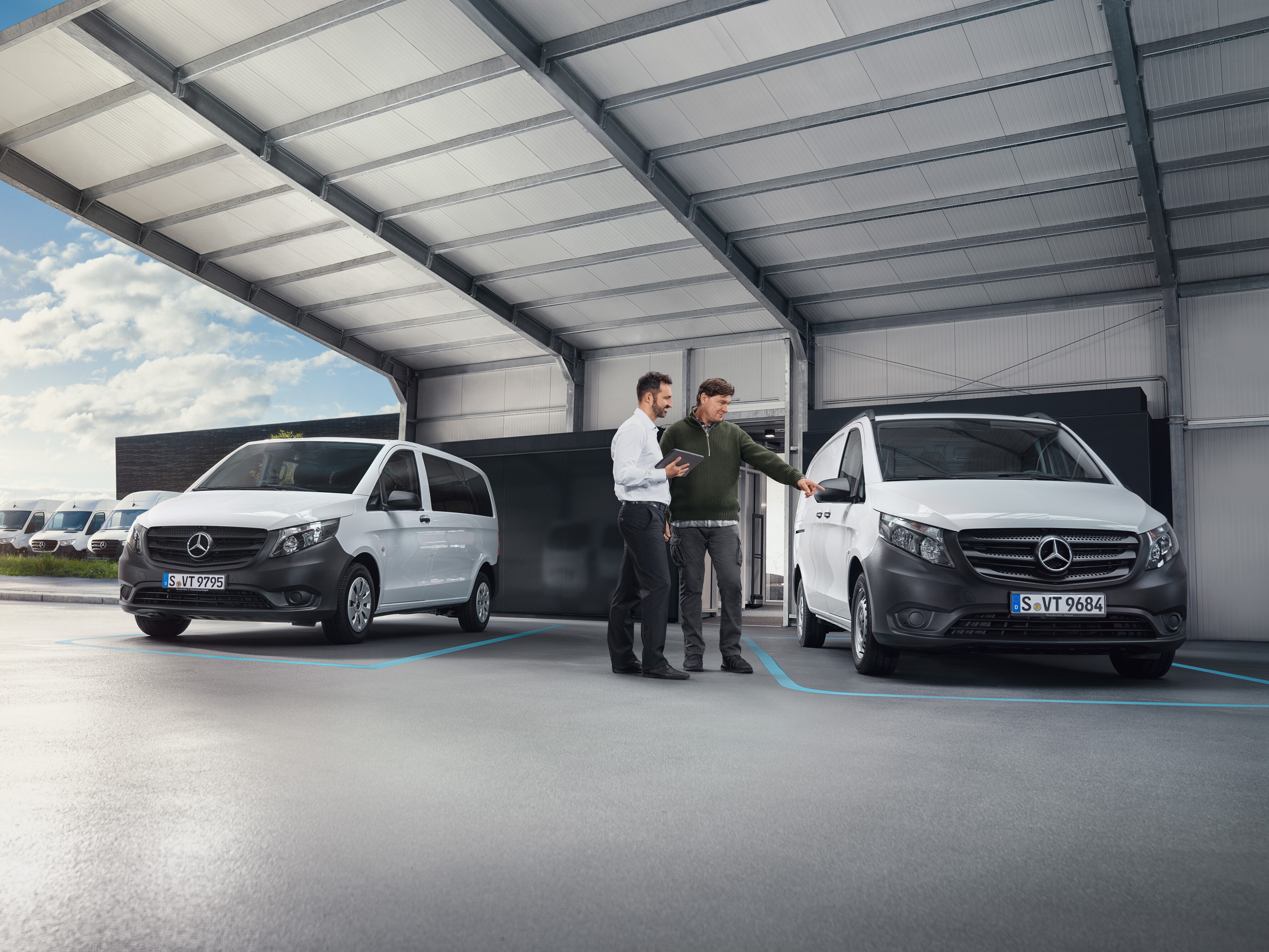 Dve stojace osoby sa rozprávajú v dielni pred dvomi úžitkovými vozidlami Mercedes-Benz.