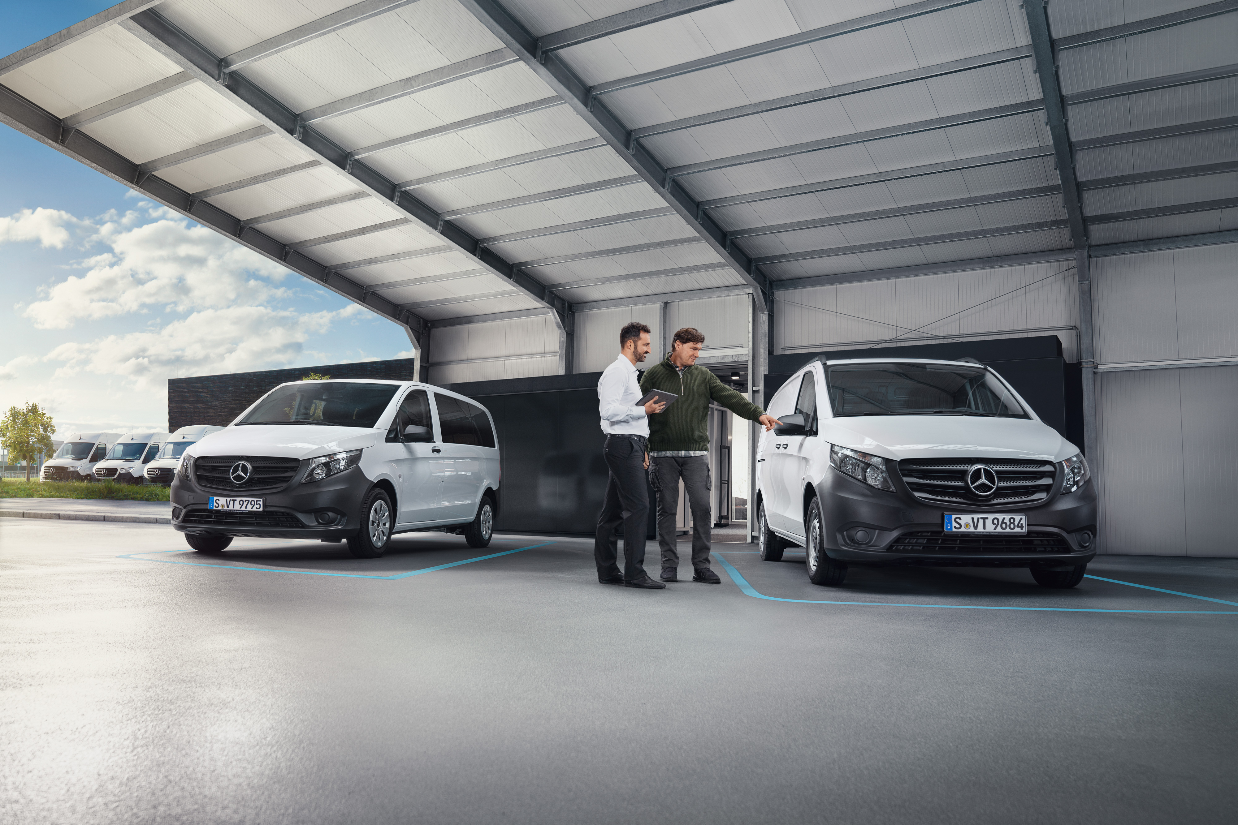 Mercedes-Benz | Pohodlie | Digitálna recepcia pre zákazníkov Dve stojace osoby sa rozprávajú v dielni pred dvomi úžitkovými vozidlami Mercedes-Benz.
