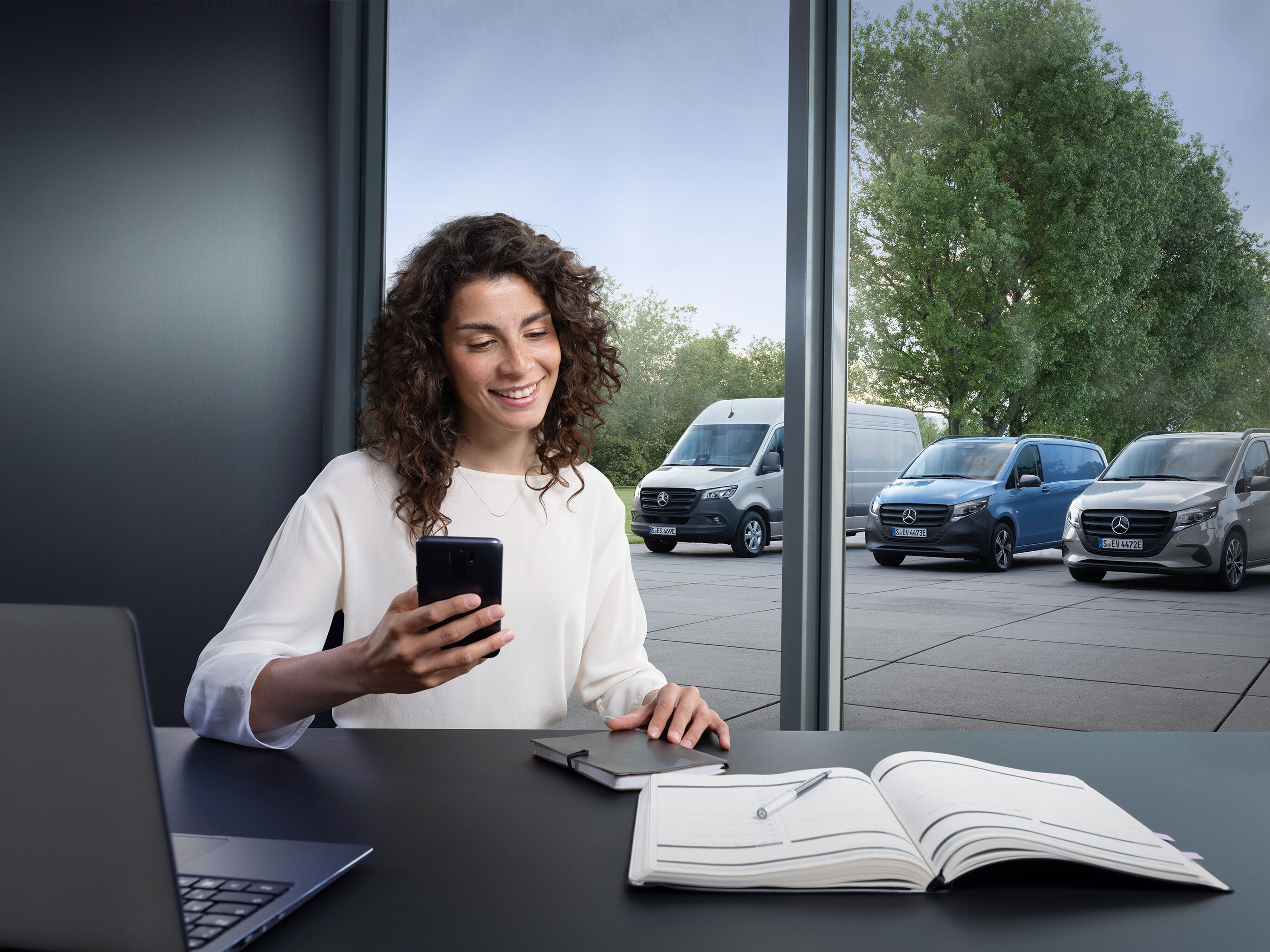 Žena si rezervuje servis prostredníctvom chytrého telefónu, v pozadí niekoľko úžitkových vozidiel Mercedes-Benz.