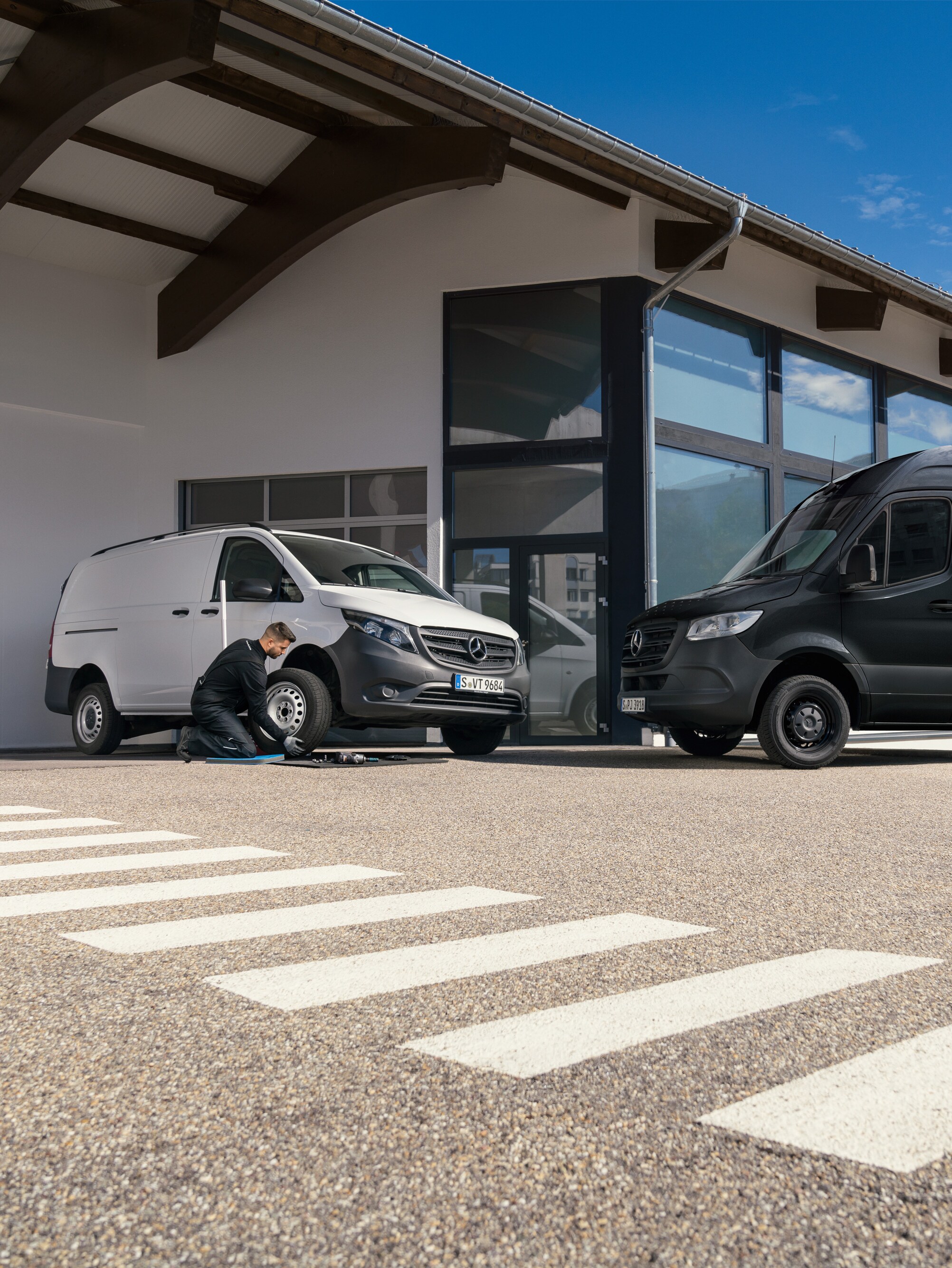 Technik vymieňa pneumatiku na bielom úžitkovom vozidle Mercedes-Benz Vito pred prezentačnou miestnosťou.