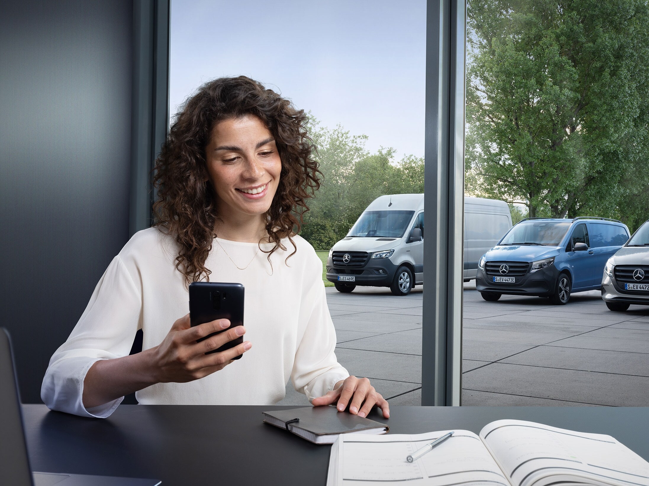 Žena používa smartfón na ovládanie monitorovania niekoľkých úžitkových vozidiel Mercedes-Benz pri okne kancelárie.